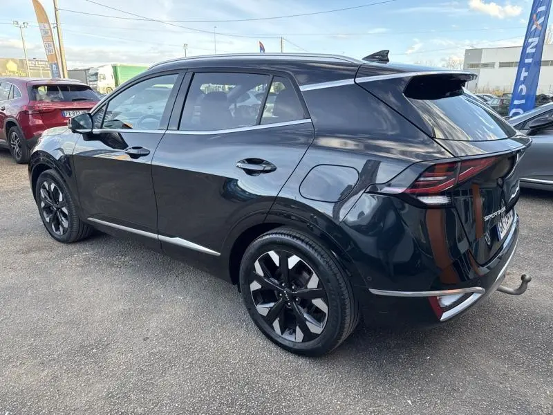 Vue 3/4 arrière droite du KIA SPORTAGE noir Basalte métallisé avec jantes alliage et attelage visible sur parking extérieur.