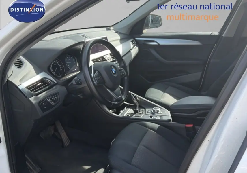 Vue intérieure côté conducteur d'un BMW X1 blanc 2022, volant multifonction et sièges tissu noirs visibles.
