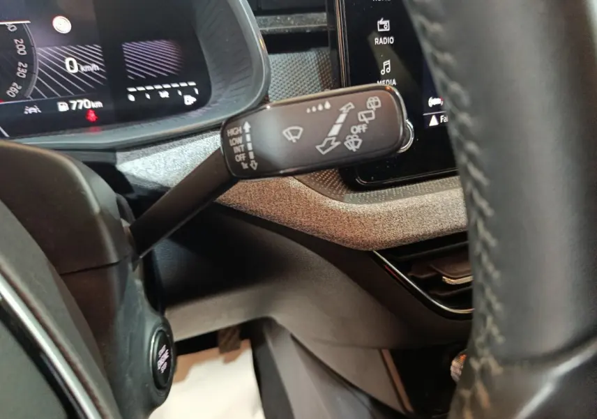 Gros plan sur la commande d’essuie-glace à gauche du volant cuir, avec tableau de bord digital et écran tactile en arrière-plan.