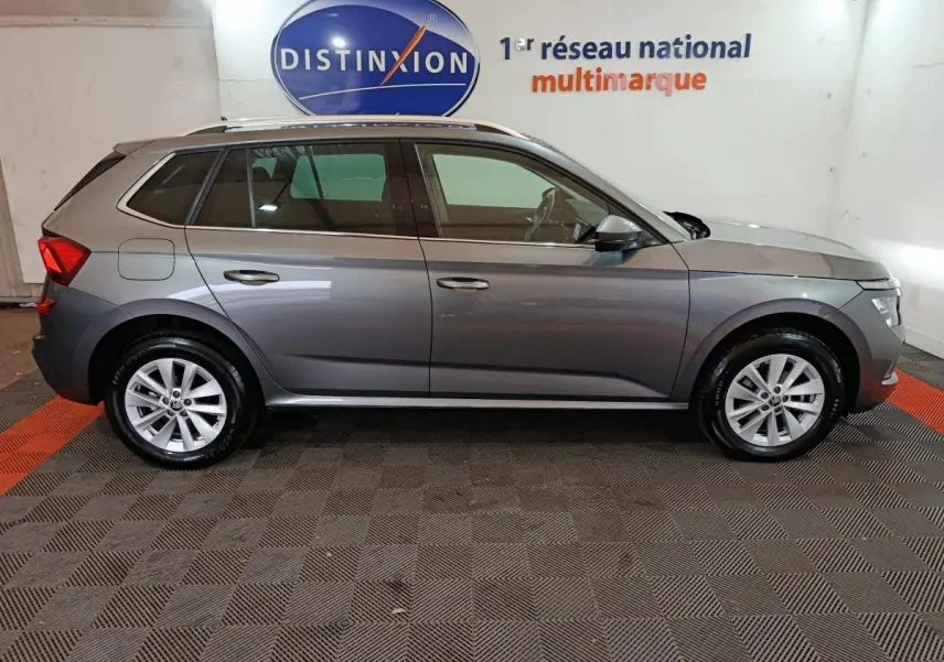 Vue de profil côté gauche d’un Skoda Kamiq gris clair 2024 garé en intérieur avec jantes alliage et barres de toit noires.