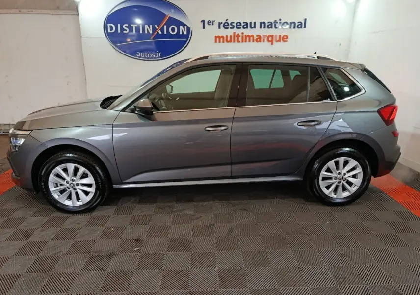 Vue de profil côté gauche d'un Skoda Kamiq gris clair 2024 avec jantes alliage et barres de toit noires.