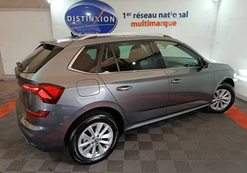 Vue 3/4 arrière droite d’un Skoda Kamiq gris clair 2024 avec jantes alliage et barres de toit noires.