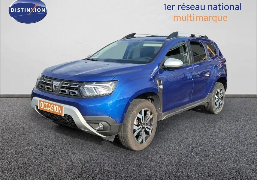 Dacia Duster bleu Iron en 3/4 avant droit avec jantes bi-ton et barres de toit visibles.