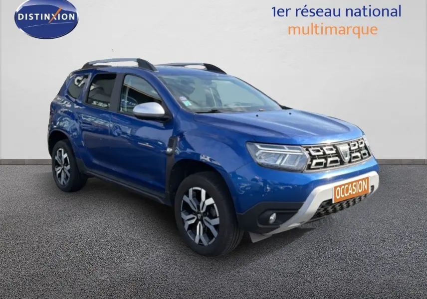 Dacia Duster bleu Iron en 3/4 avant droit, SUV compact avec jantes bi-ton et barres de toit visibles.