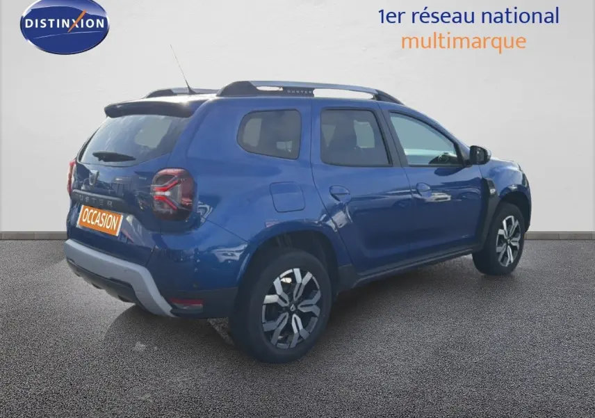 Vue 3/4 arrière droite d'un Dacia Duster 1.0 ECO-G 100 Prestige bleu Iron avec jantes bi-ton et barres de toit.