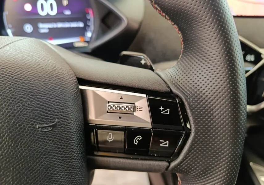 Gros plan sur le volant cuir perforé du DS3 2024 avec commandes vocales et téléphone, combiné numérique flou en arrière-plan.