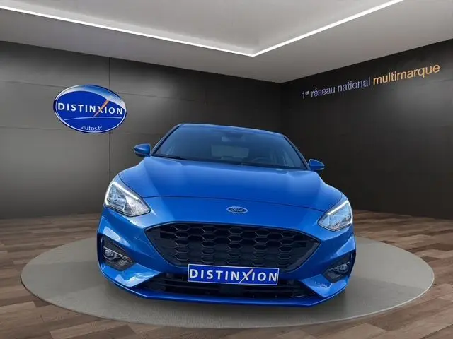 Vue frontale d'une Ford Focus 1.5 EcoBoost 150ch ST-Line BVA bleue, avec calandre noire et phares LED allumés.
