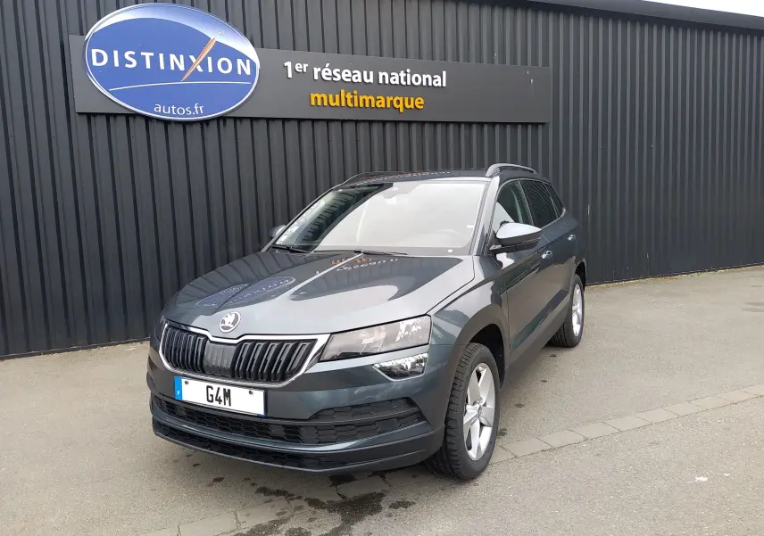 Skoda Karoq gris en 3/4 avant droit, SUV compact avec calandre noire et jantes argentées, stationné devant un mur noir.