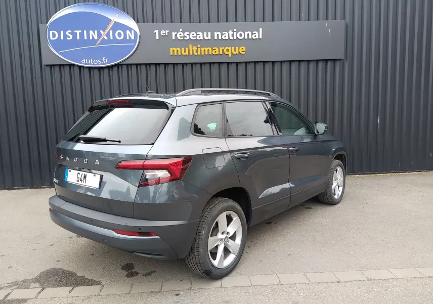 Vue 3/4 arrière droite d'un Skoda Karoq gris 2021 garé devant un mur noir avec un panneau Distinxion.