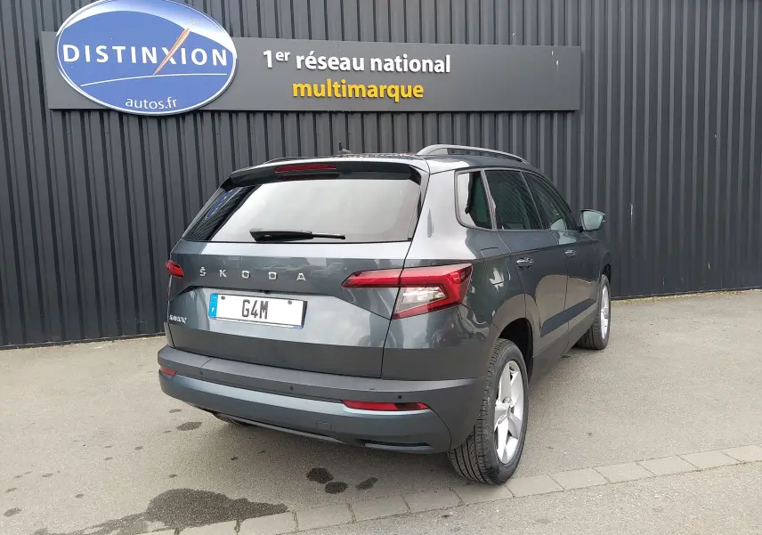 Vue 3/4 arrière droite d'un Skoda Karoq gris 2021 avec jantes alliage et feux arrière LED, stationné devant un mur noir.