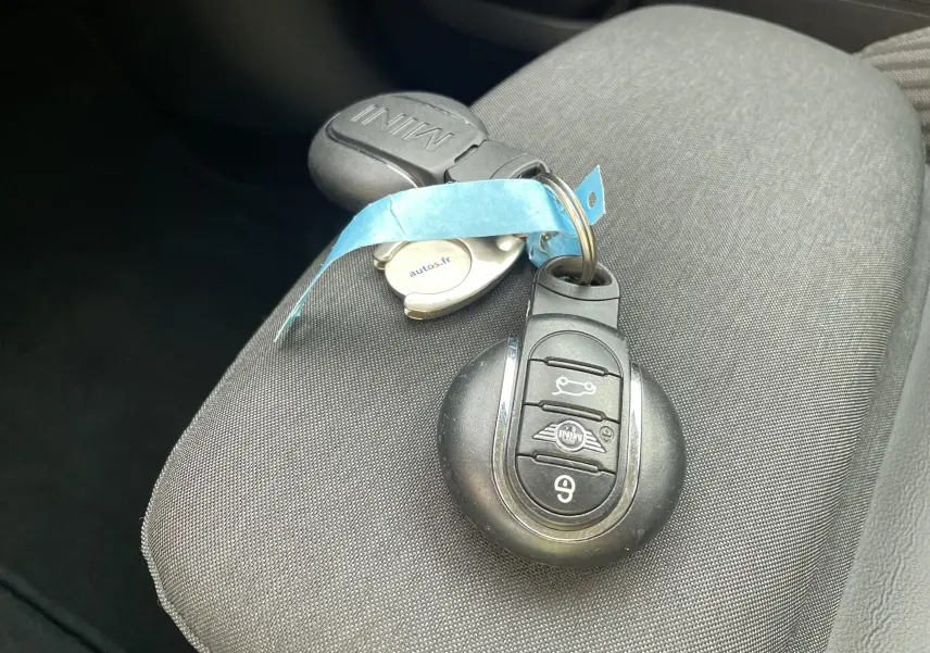 Gros plan sur la clé MINI posée sur un siège gris foncé, avec un porte-clé bleu et un badge autos.fr visible.
