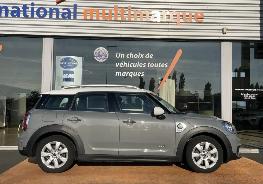 Profil côté droit d'une MINI COUNTRYMAN F60 hybride rechargeable Moonwalk Grey garée devant une vitrine multimarque.