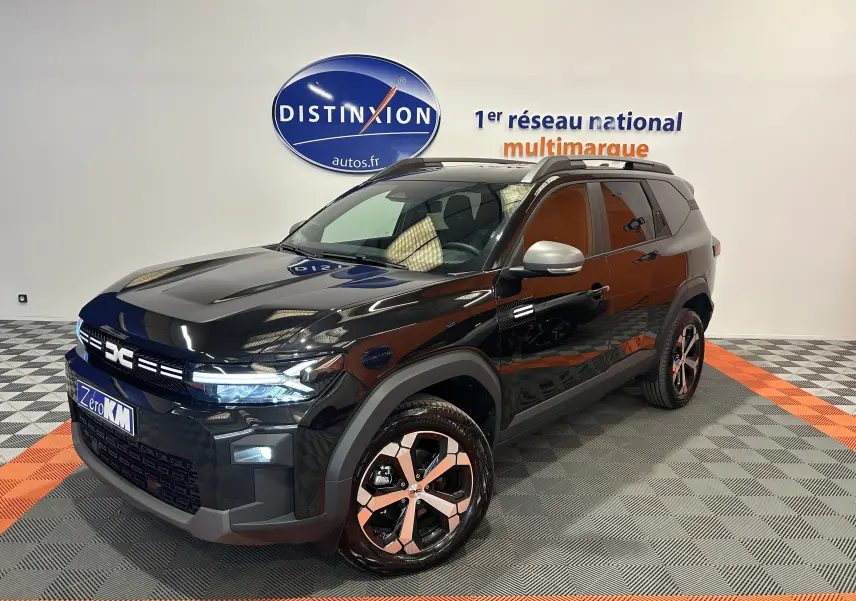 Dacia Bigster 2026 noir nacré en 3/4 avant droit, avec jantes bicolores et hayon électrique visible en arrière-plan.