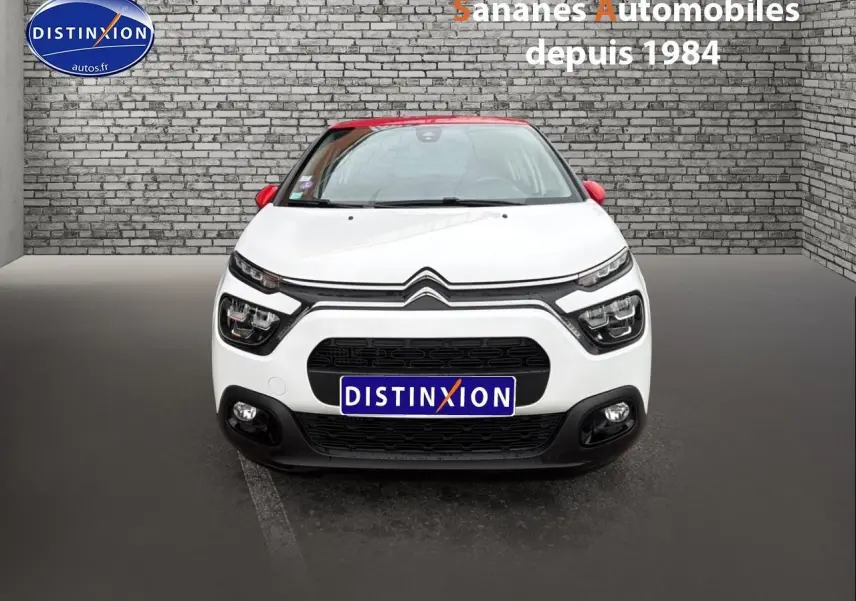 Vue frontale d'une Citroën C3 blanche avec toit et rétroviseurs rouges, sur fond de mur en briques grises.