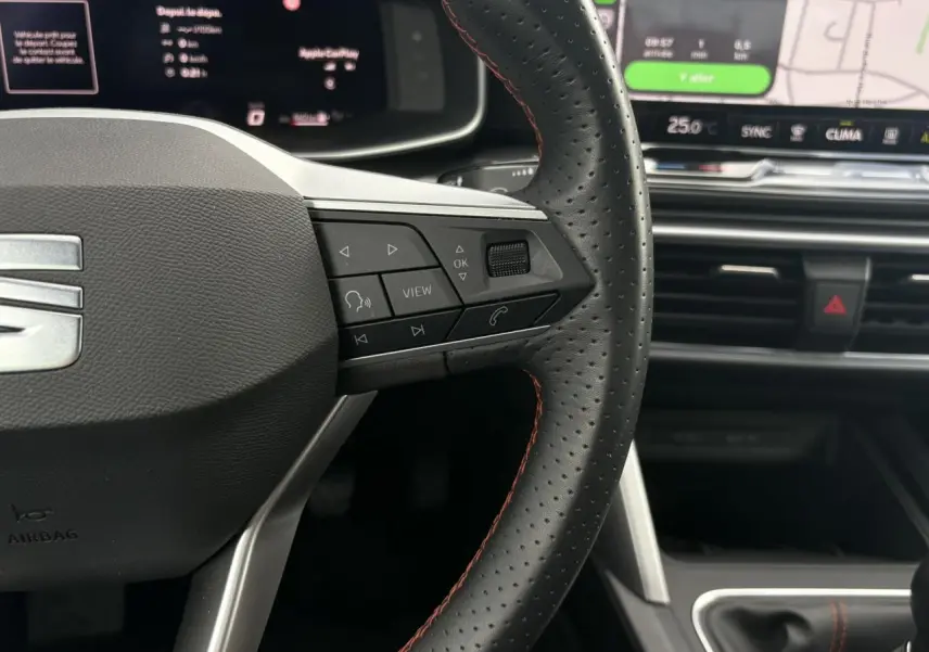 Gros plan sur le volant en cuir noir perforé avec surpiqûres rouges du SEAT Leon 2025, tableau de bord et écran tactile en arrière-plan.