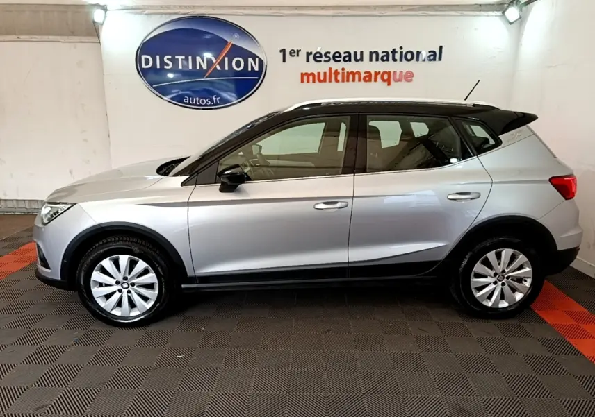 Vue de profil côté gauche d’un SEAT Arona gris clair avec toit noir et jantes alliage en intérieur showroom.