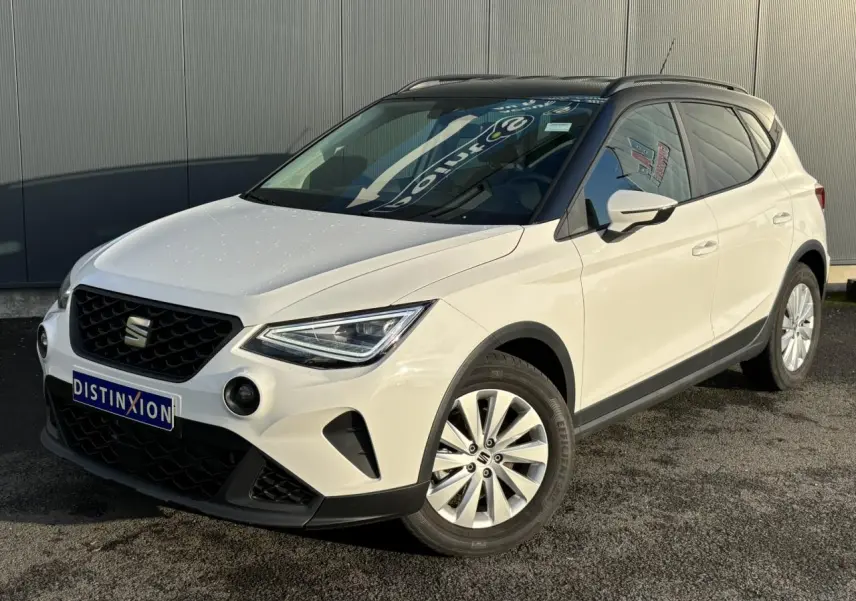 Vue 3/4 avant droite d'un SEAT Arona blanc avec toit noir et jantes alliage 16 pouces, stationné devant un mur gris.