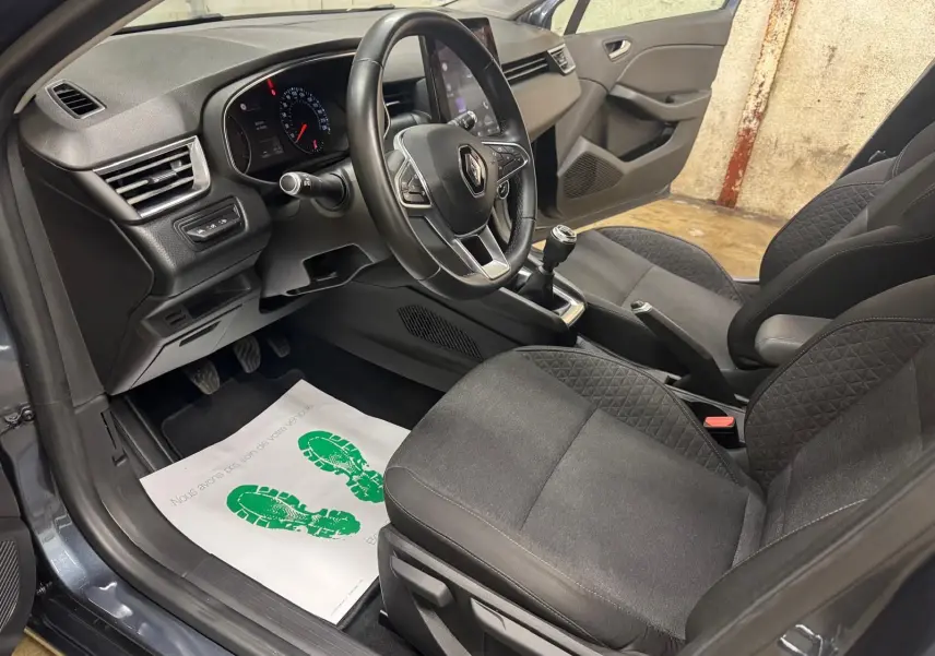 Vue intérieure côté conducteur de la Renault Clio V Business gris titanium, montrant le volant, la boîte manuelle et les sièges en tissu noir.