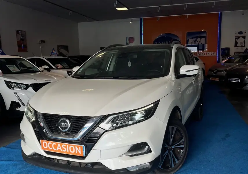 Nissan Qashqai blanc lunaire en 3/4 avant droit, avec jantes noires et plaque "OCCASION" en showroom.