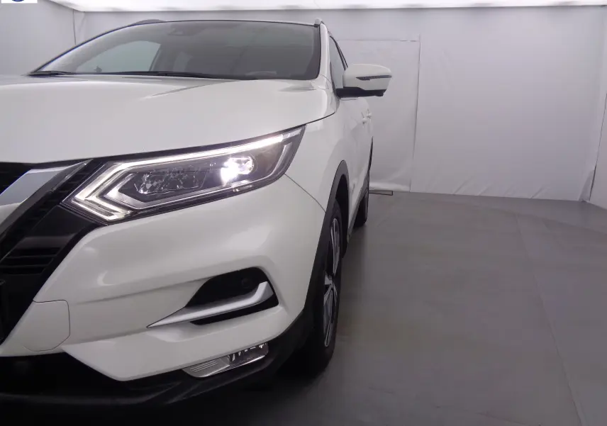 Vue 3/4 avant droit du Nissan Qashqai blanc lunaire métal avec phares allumés dans un studio lumineux.