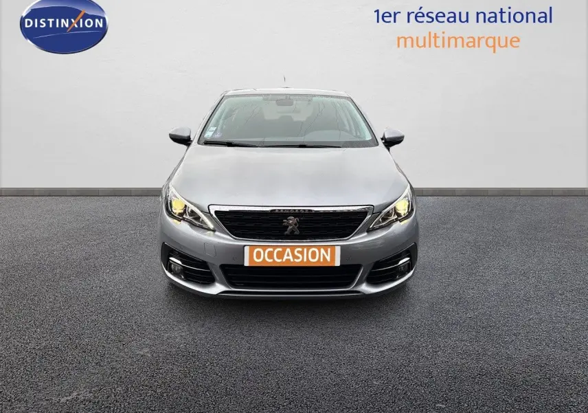 Vue frontale d'une Peugeot 308 gris artense avec phares allumés et plaque d'immatriculation orange "OCCASION" visible.