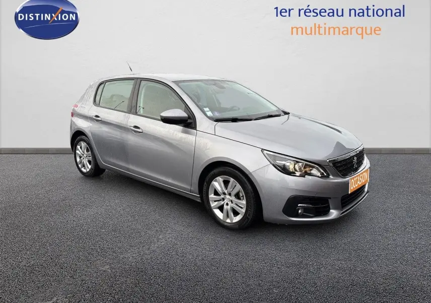 Peugeot 308 gris Artense vue 3/4 avant droit, compacte cinq portes avec feux allumés sur fond neutre.