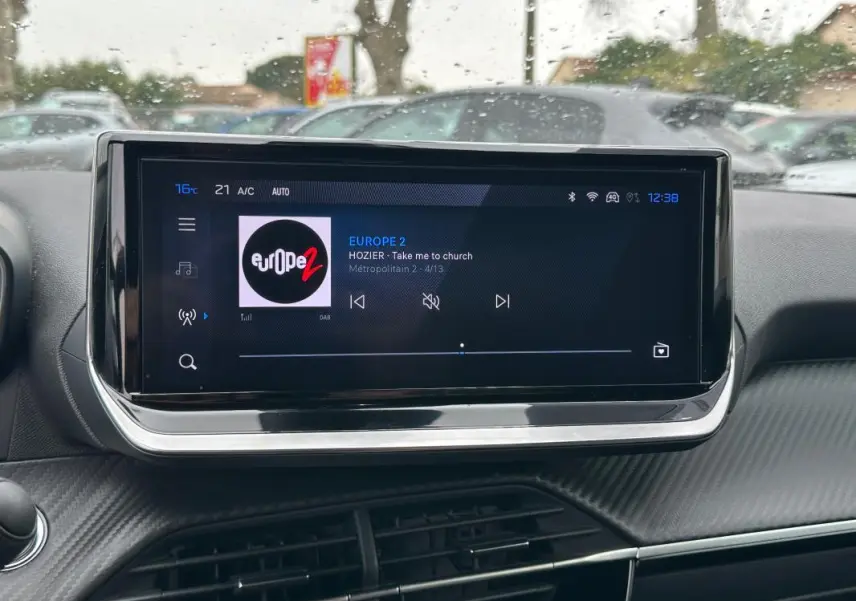 Écran tactile central du tableau de bord du Peugeot 2008 Hybrid 145 Allure, affichant une radio en extérieur pluvieux.