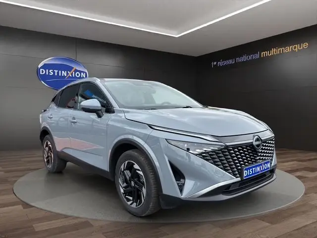 Vue 3/4 avant du Nissan Qashqai 2025 gris céramique avec hayon motorisé et calandre distinctive en showroom.