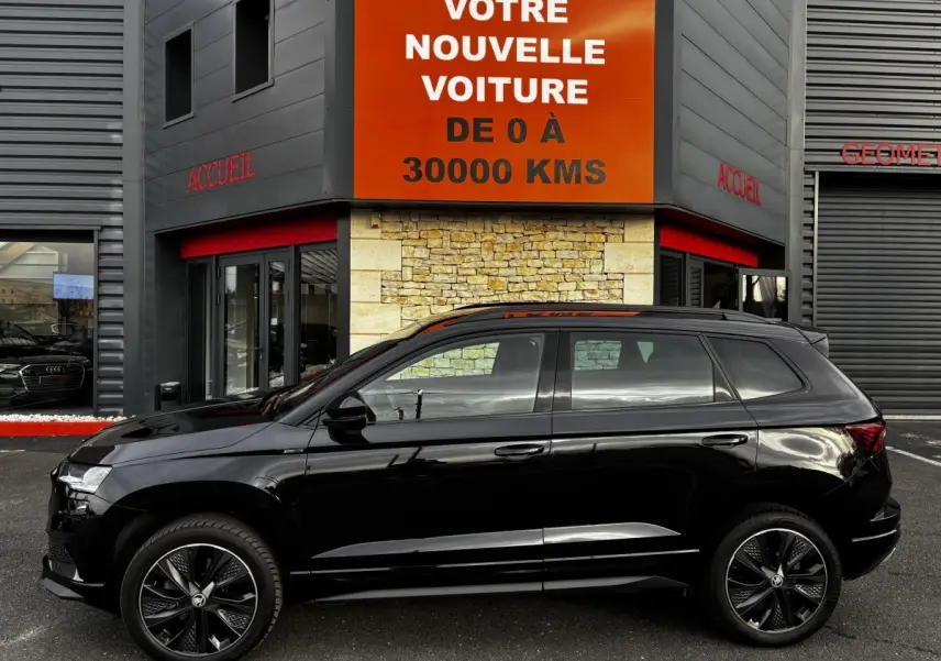 Profil côté gauche d'un SUV SKODA KAROQ noir 2025 garé devant un bâtiment avec panneau orange "Votre nouvelle voiture".
