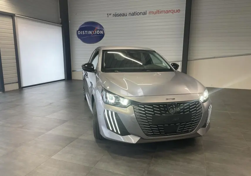 Peugeot 208 gris Artense en 3/4 avant droit, avec calandre noire et feux LED allumés dans un showroom.