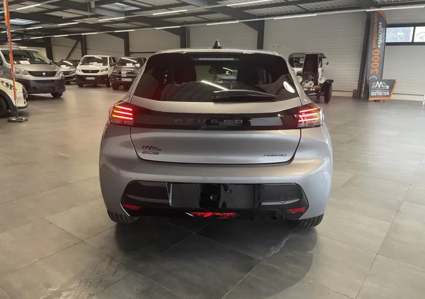 Vue arrière d'une Peugeot 208 hybride gris Artense avec feux arrière LED allumés en intérieur showroom.