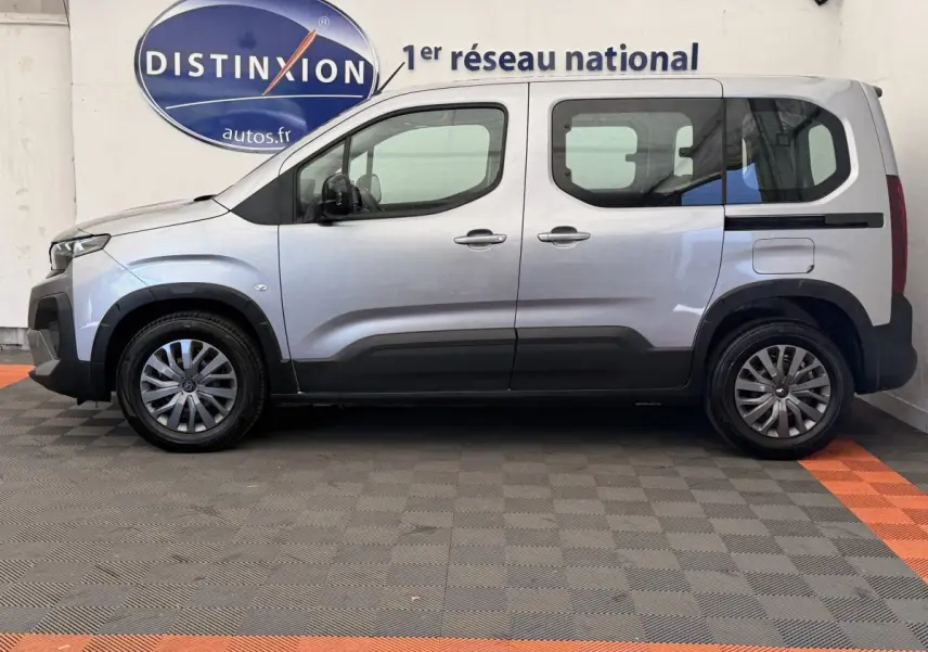 Vue de profil côté gauche d'un Peugeot Rifter gris clair 2024 stationné en intérieur sur sol damier.