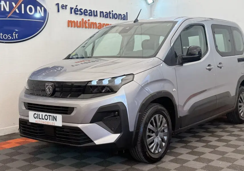 Peugeot Rifter gris clair en 3/4 avant droit, avec calandre noire et feux LED distinctifs, stationné en intérieur.