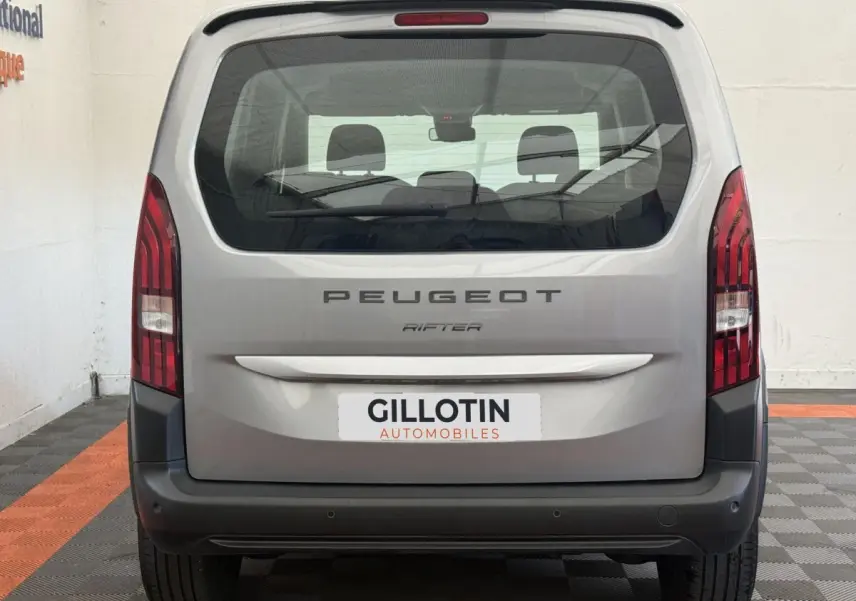 Vue arrière du Peugeot Rifter gris clair 2024 avec feux verticaux et pare-chocs noir mat visible en intérieur.