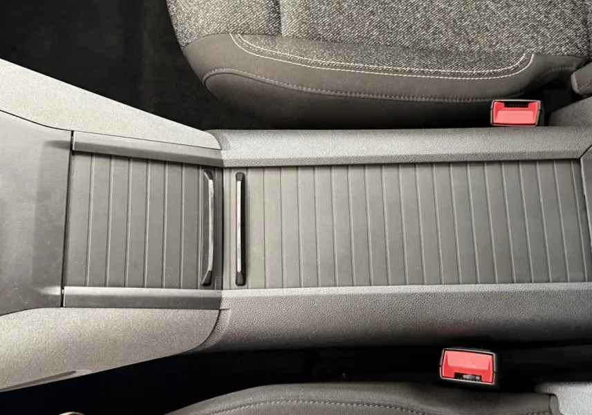 Gros plan sur la console centrale grise du Peugeot Rifter 2024, avec sièges tissu et boucles de ceinture rouges visibles.