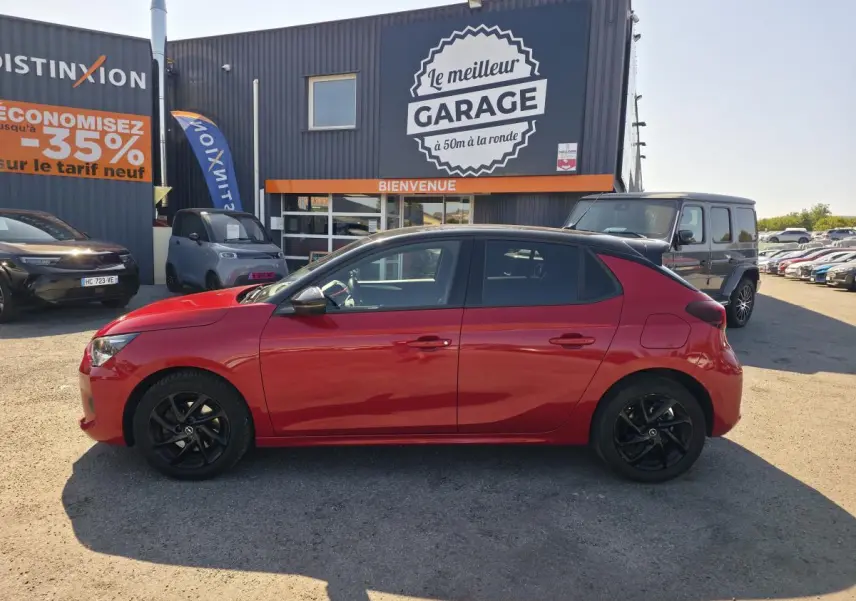 Opel Corsa rouge vue de profil côté gauche, avec toit noir et jantes alliage noires, stationnée devant un garage.