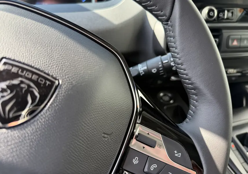 Gros plan sur le volant cuir noir du Peugeot Rifter gris acier, avec commandes intégrées et logo lion central.