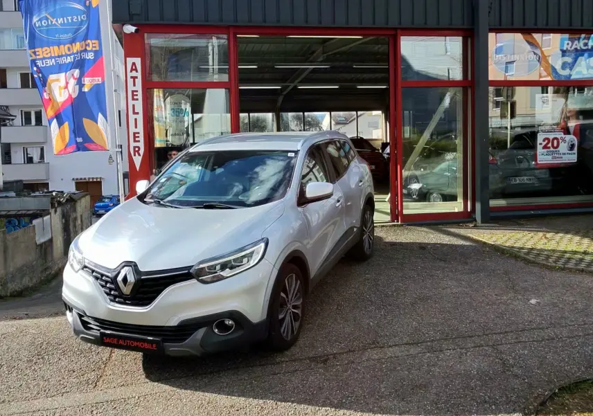Renault Kadjar gris platine métal vu en 3/4 avant droit devant un garage AD Expert avec enseigne rouge et blanche.
