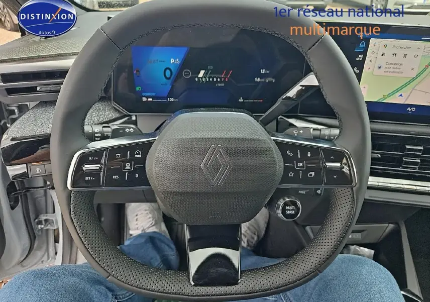 Vue intérieure centrée sur le volant noir perforé de la Renault Clio VI avec tableau de bord numérique et écran tactile.