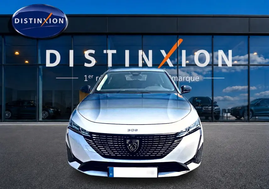 Vue frontale d'une Peugeot 308 SW blanche 2025 avec calandre noire et logo lion distinctif, devant un bâtiment vitré.