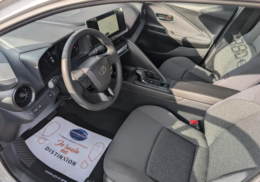 Vue intérieure côté conducteur du Toyota C-HR 2024, sellerie tissu gris foncé et tableau de bord avec écran tactile.