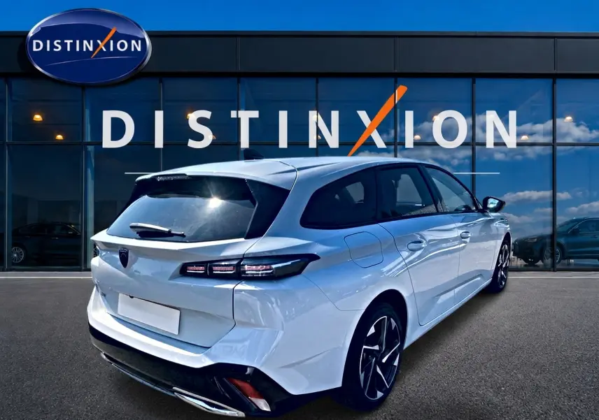 Vue 3/4 arrière droite d'une Peugeot 308 SW blanche 2025 avec jantes alu et vitres arrière surteintées, devant un showroom Distinxion.