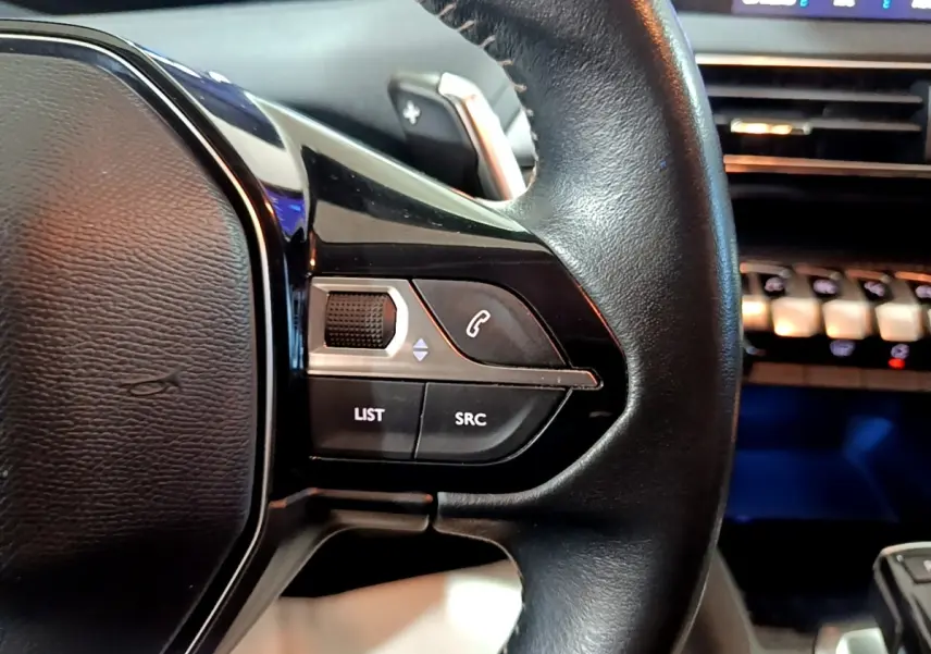 Gros plan sur la partie gauche du volant cuir noir du Peugeot 5008, avec commandes audio et téléphone intégrées.