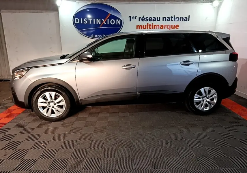 Vue de profil côté gauche d'un Peugeot 5008 gris clair 2020 stationné en intérieur avec logo Distinxion en arrière-plan.