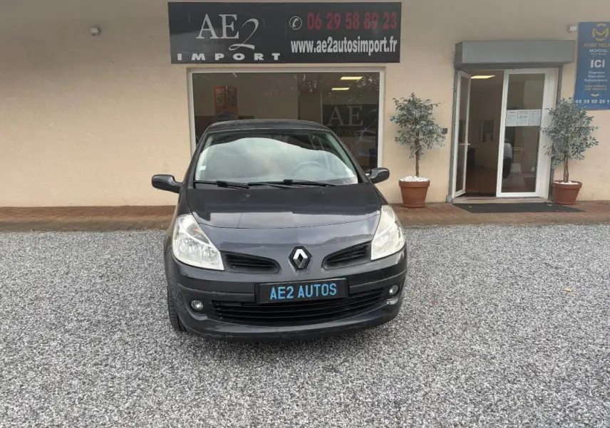 Vue avant d'une Renault Clio III gris foncé, avec plaque AE2 AUTOS, stationnée devant un bâtiment commercial.