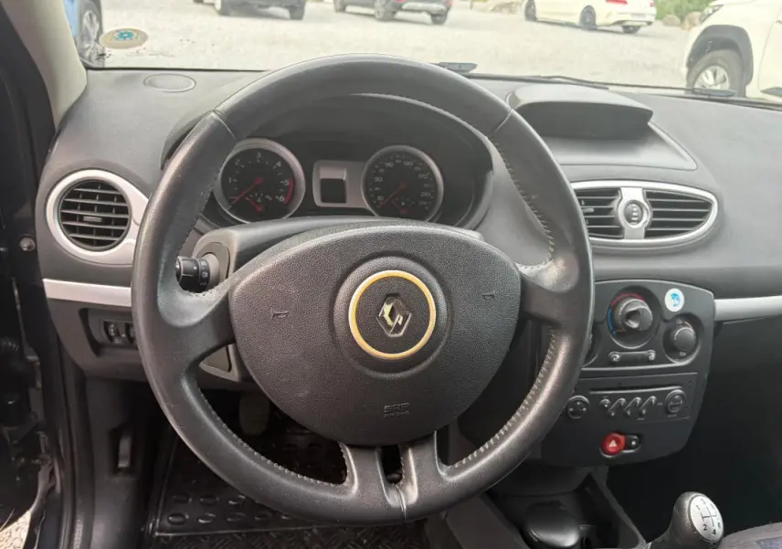 Vue intérieure centrée sur le volant noir de la Renault Clio III gris, avec tableau de bord et levier de vitesse manuel visibles.