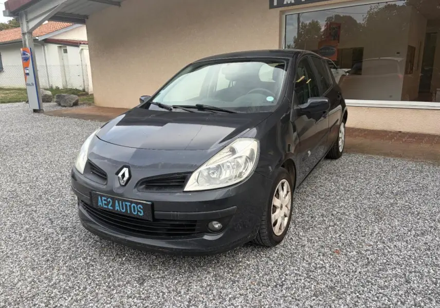 Renault Clio III gris vue 3/4 avant droit, avec plaque AE2 AUTOS, garée sur gravier devant un bâtiment.