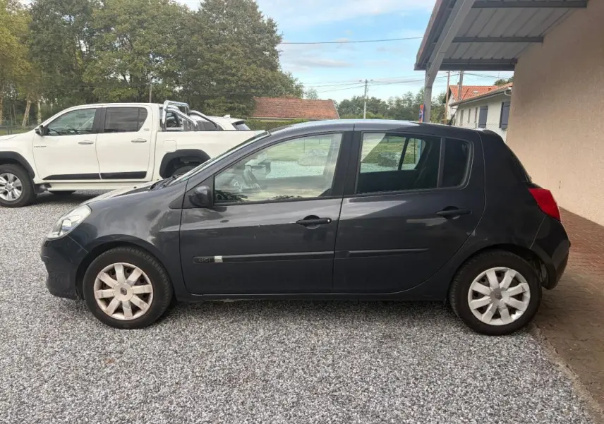 Renault Clio III gris vue de profil côté gauche, garée sur gravier devant un abri et un mur beige.