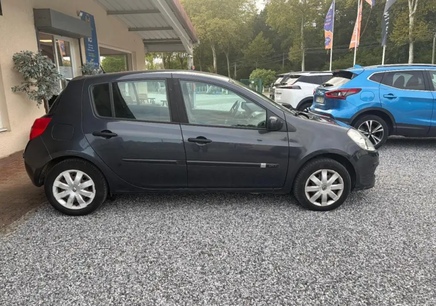 Profil côté droit d'une Renault Clio III gris foncé de 2005, garée sur un sol gravillonné devant un bâtiment.