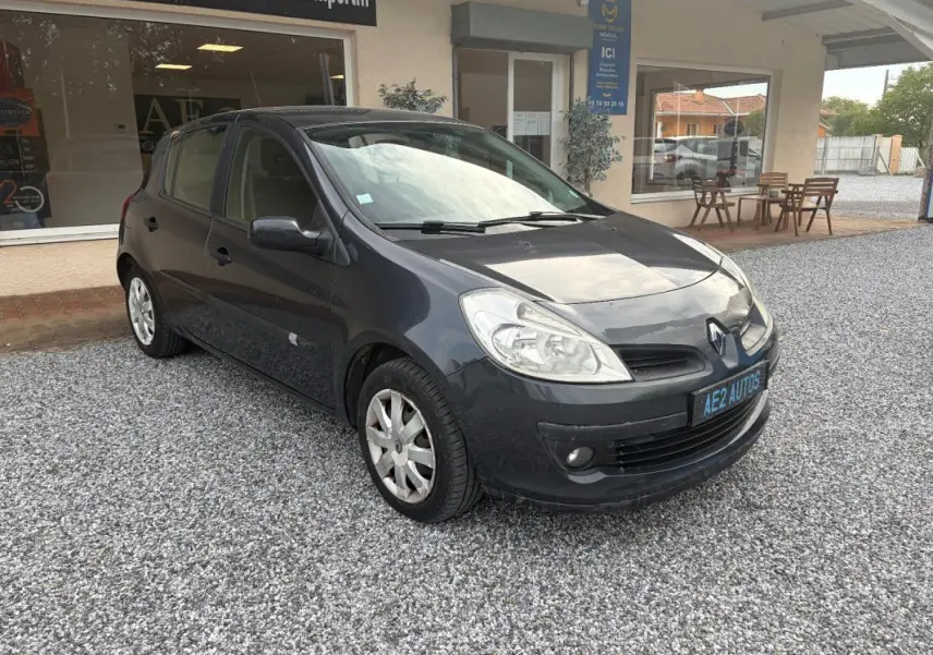 Renault Clio III gris vue 3/4 avant droit, garée sur gravier devant un bâtiment avec vitrine et terrasse.